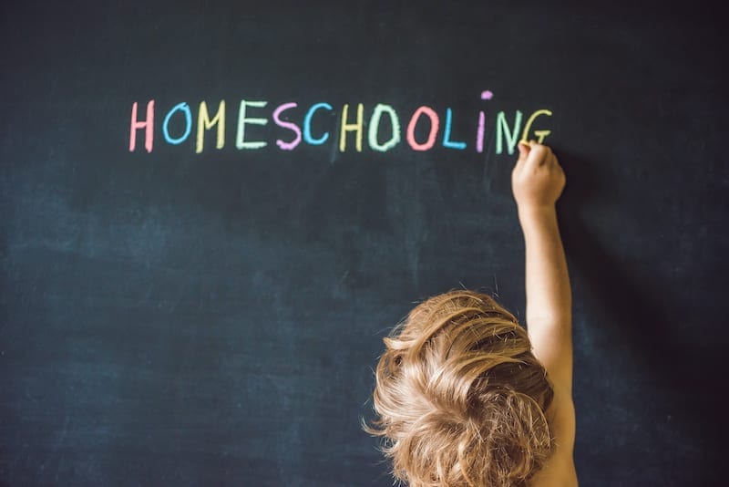 a young child writes the word "homeschooling" in many different colors on a chalkboard