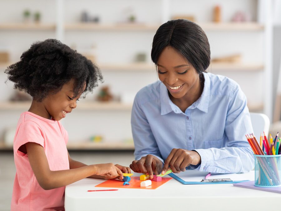 Adorable little black girl with curly hair having fun at daycare, happy kid and child development specialist attractive young woman cutting play dough on board, arts and crafts with children