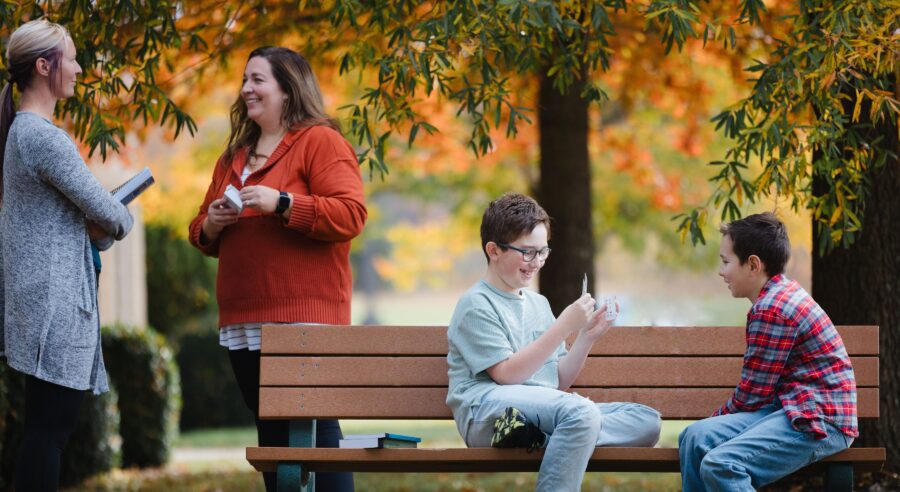 two homeschool moms talking while two students learn with math flashcards on a park bench
