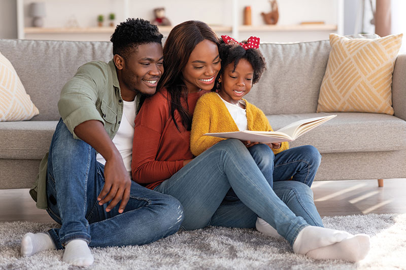 Mother and Father sitting on the floor with their daughter while she is reading at a book