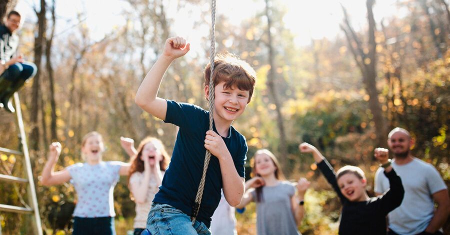 Boy in blue shirt playing on a rope swing with friends cheering in the background