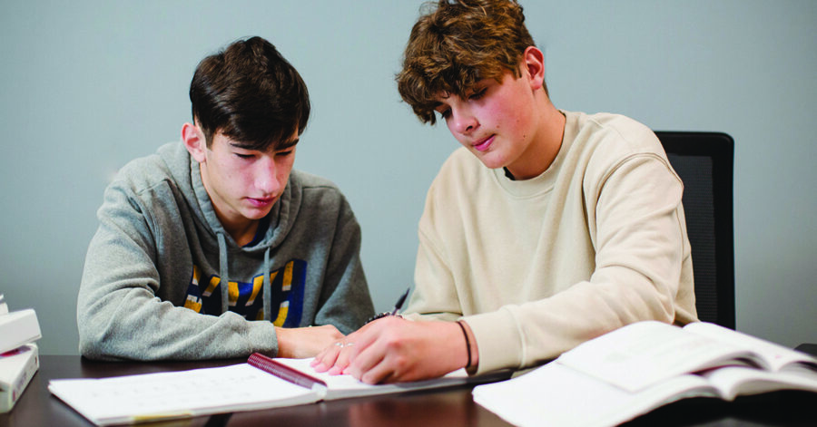 Two teenage boys working together at a table, writing and reviewing notes, illustrating the process of organizing academic records for a homeschool transcript.